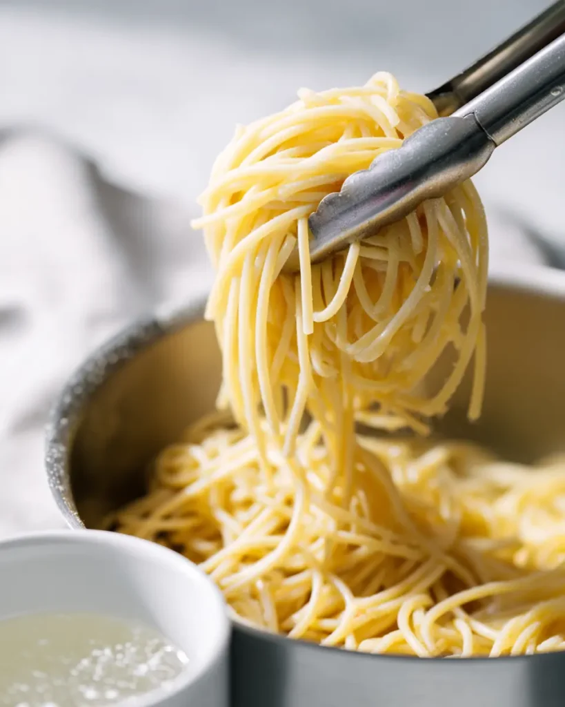 Ultimate Spaghetti Carbonara Recipe (Creamy, No Cream) 14 Al dente spaghetti being lifted from a pot with tongs and starchy pasta water reserved in a white cup for the carbonara recipe.